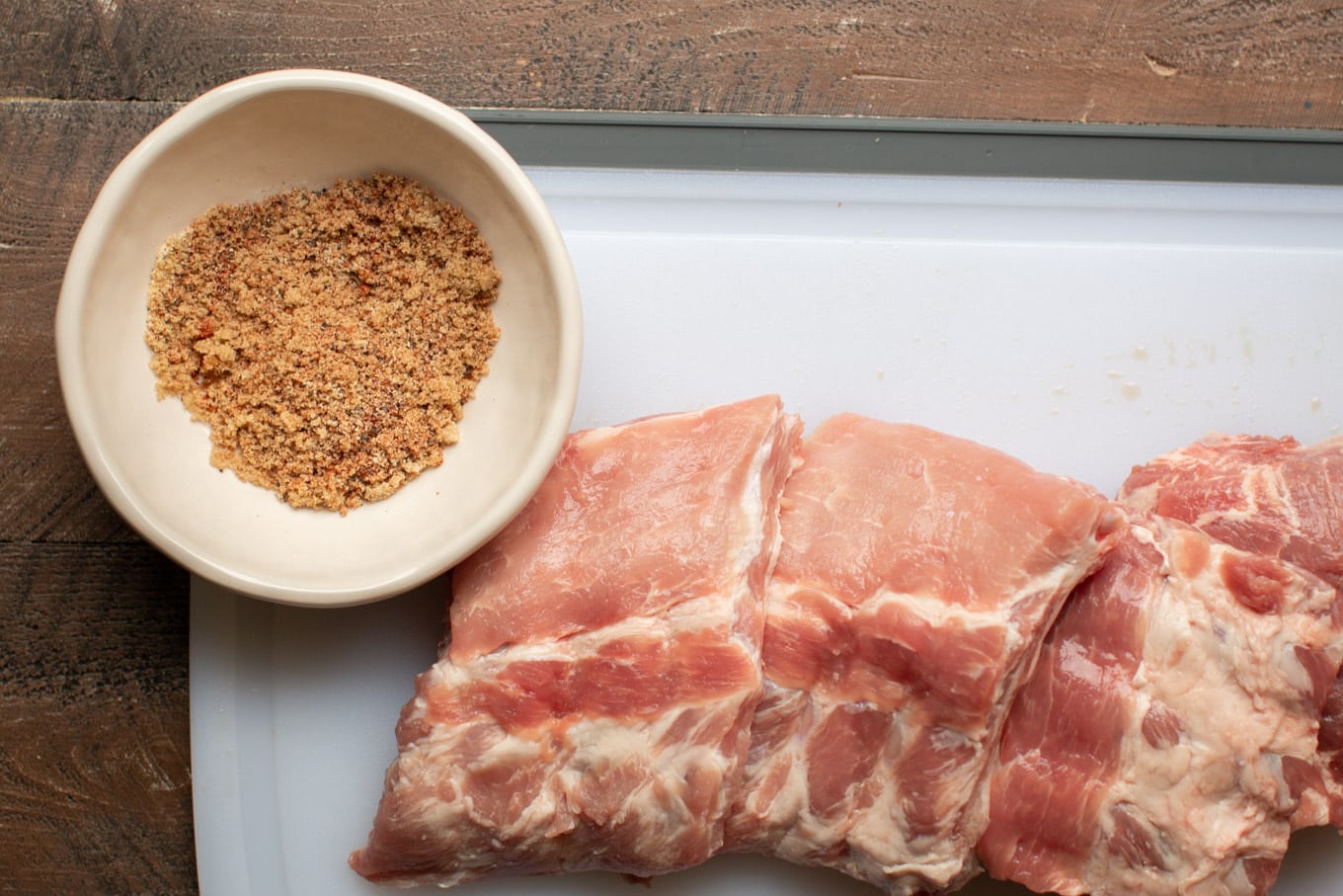 Raw rack of ribs, cut into 4 sections with bowl of rub next to it.