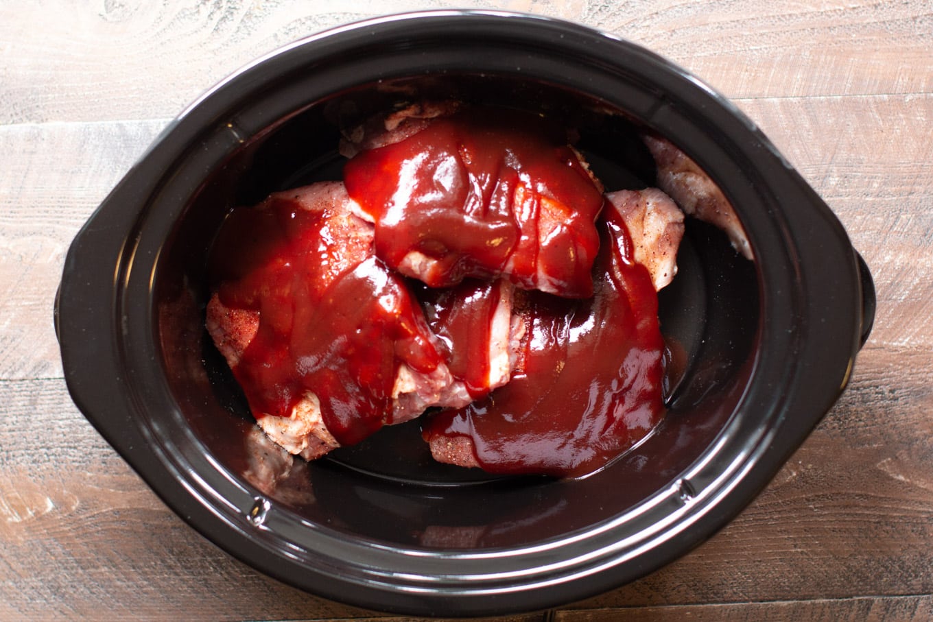 Raw ribs in the slow cooker with barbecue sauce on top.