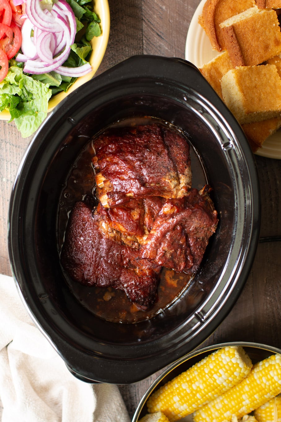 4 sections of barbecue ribs in the slow cooker with corn, salad and cornbread on the side.