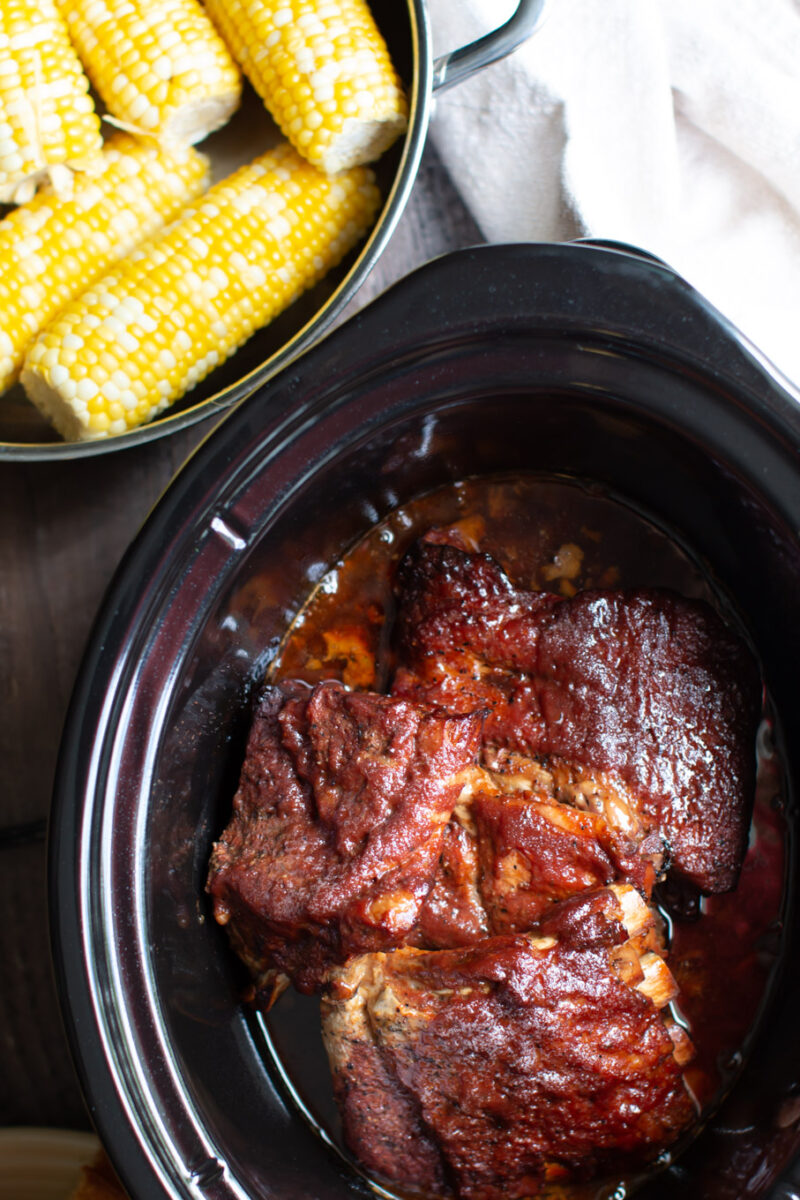 Cooked barbecue ribs in the slow cooker with corn on the cob on the side.