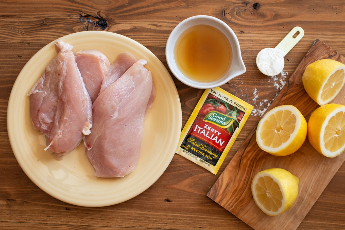 chicken breasts, chicken broth, zesty Italian seasoning packet, cornstarch and lemons on wood table.