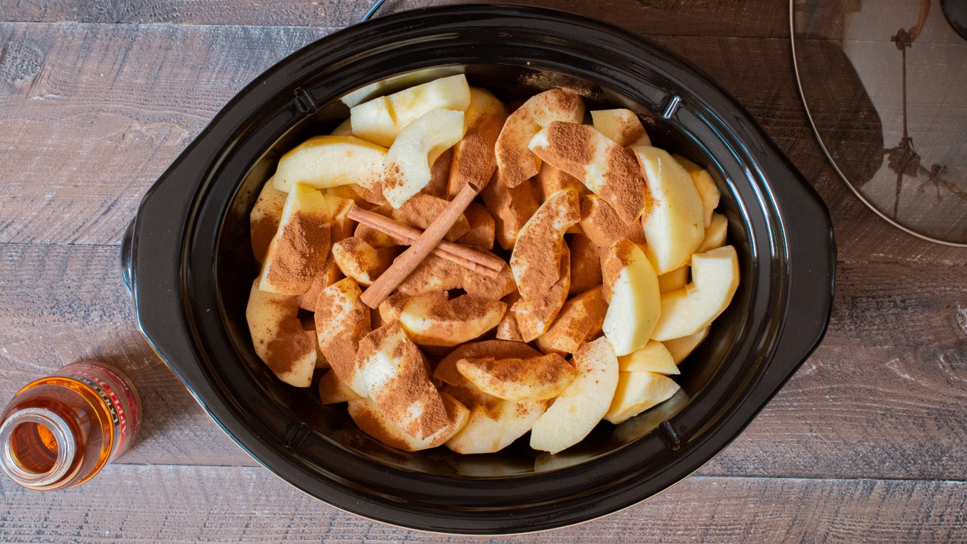 Sliced apples with powdered cinnamon and cinnamon sticks on top in a slow cooker.