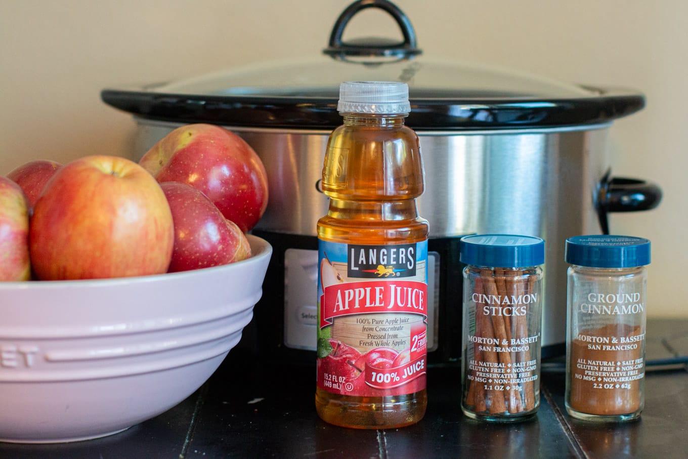 Apples, apple juice, cinnamon and cinnamon sticks in front of a slow cooker.