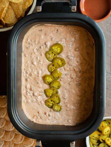 Cooked taco dip in slow cooker with jalapenos on top, chips and jalapenos on side.