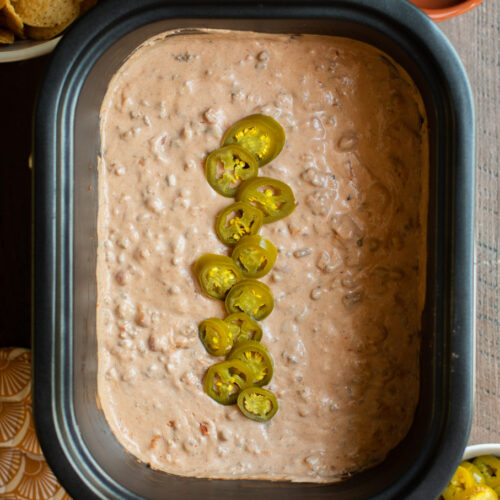 Cooked taco dip in slow cooker with jalapenos on top, chips and jalapenos on side.