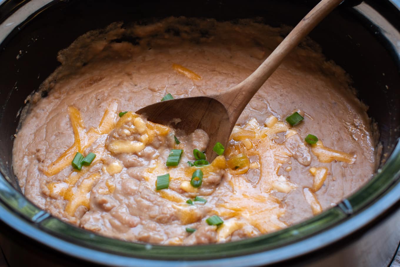 close up of refried beans in a slow cooker with wooden spoon in it.