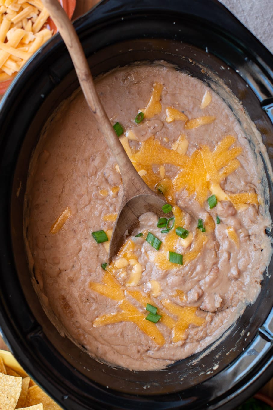 refried beans in a slow cooker with cheddar cheese and green onions on top.