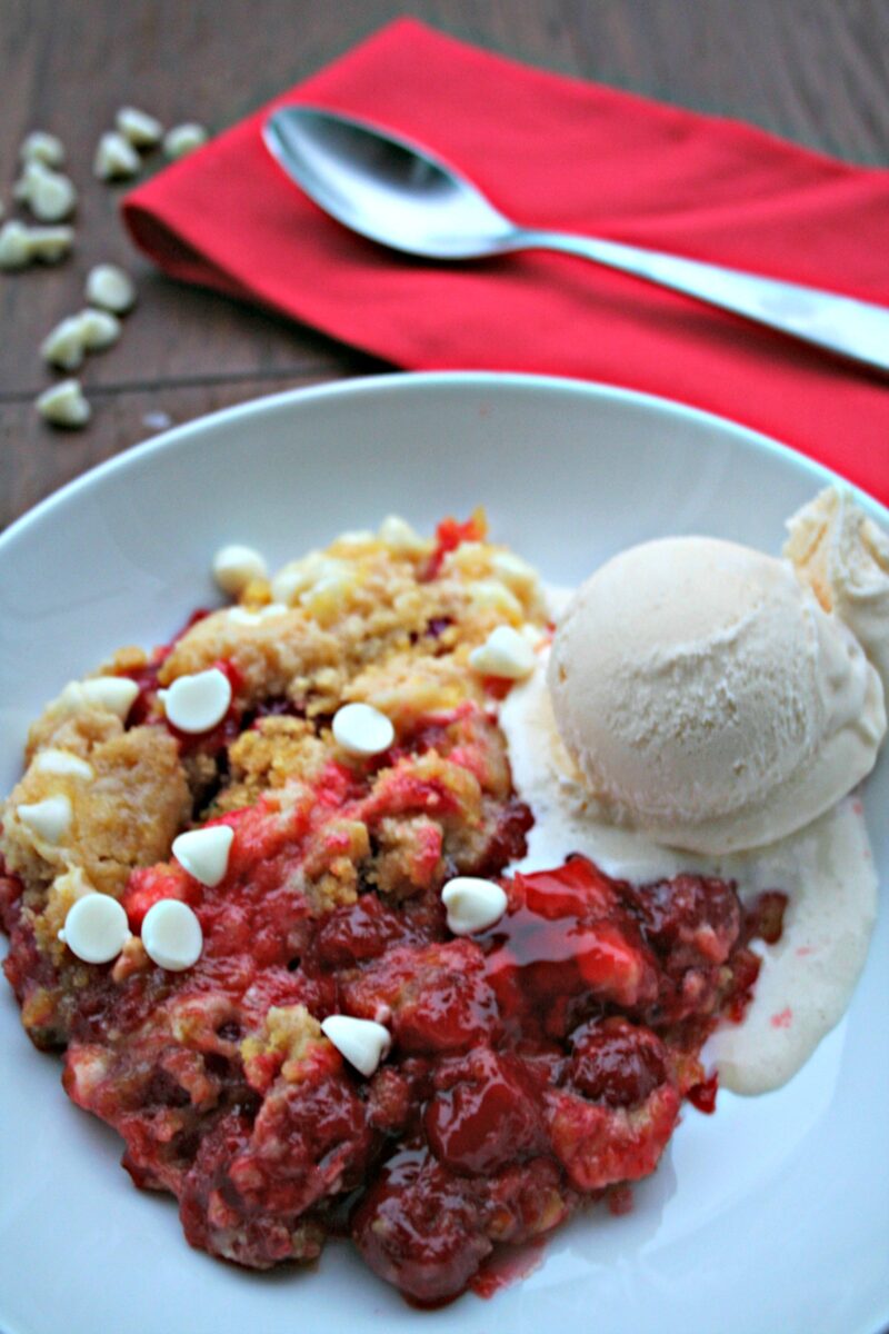 close up of white chocolate cherry dump cake in a white bowl with ice cream on top