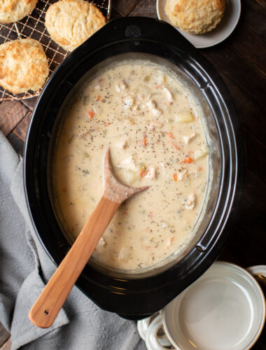 Chicken pot pie filling in slow cooker with spoon in it and biscuits on side