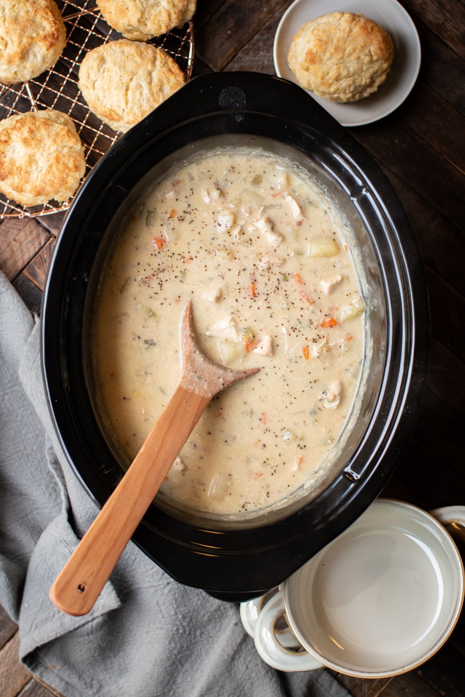Chicken pot pie filling in slow cooker with spoon in it and biscuits on side