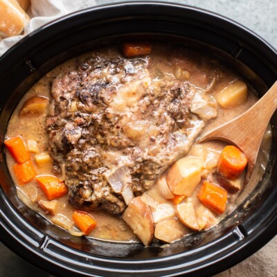 pot roast with gravy, potatoes and onions in a slow cooker with wooden spoon in it.