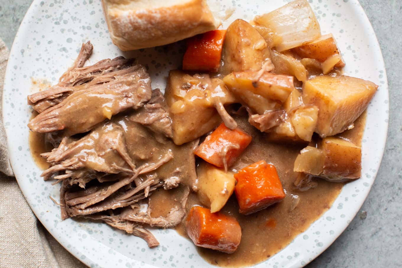 pot roast with vegetables dinner roll and gravy on a speckled plate.