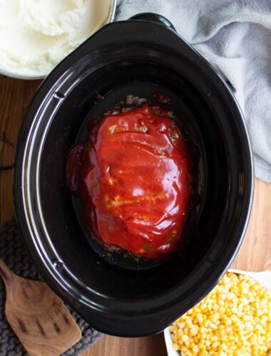 Meatloaf in a slow cooker with mashed potatoes and corn on the side.