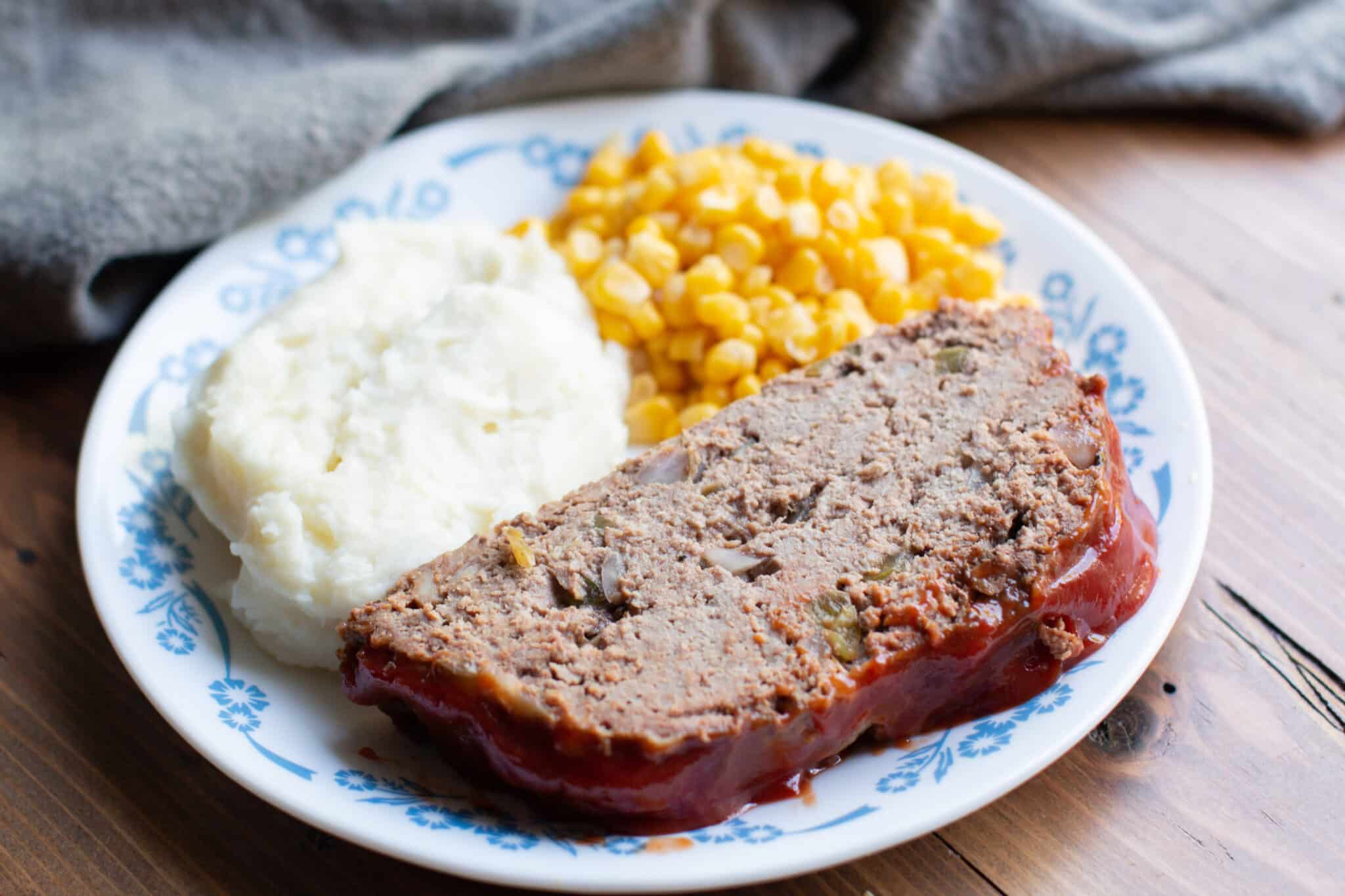 sliced cooked meatloaf on a white plate with mashed potatoes and corn.