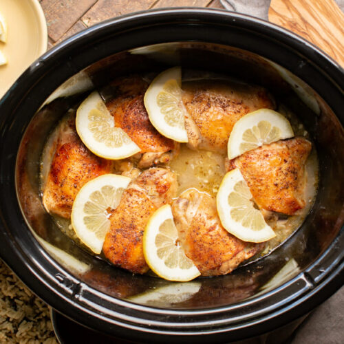 cooked lemon pepper chicken thighs in slow cooker with lemon slices on top.