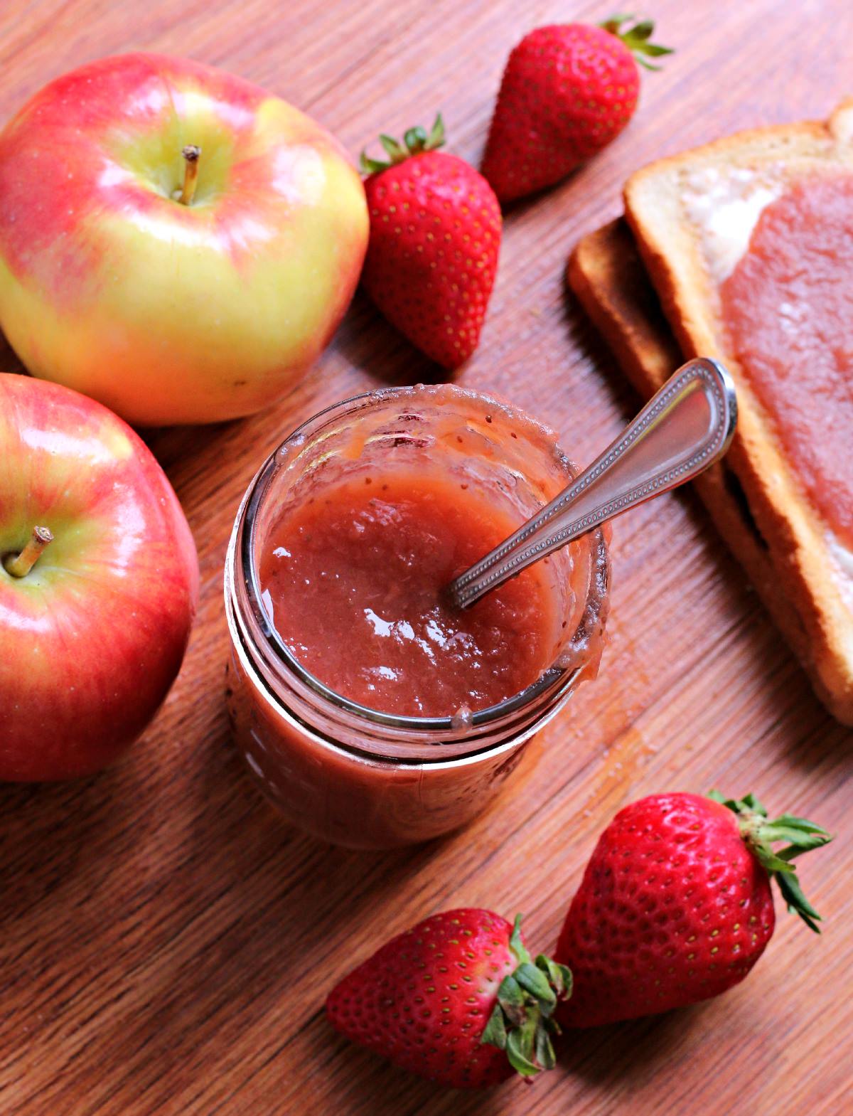 jar of strawberry applesauce with spoon in it, apples and strawberries on the side.