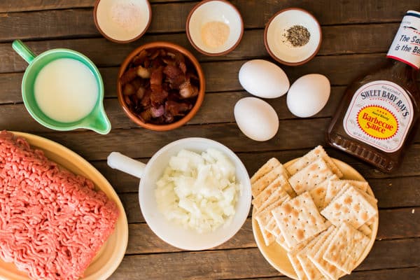 ground beef, milk, bacon, salt, pepper, eggs, crackers onion, and barbecue sauce on a wooden table.