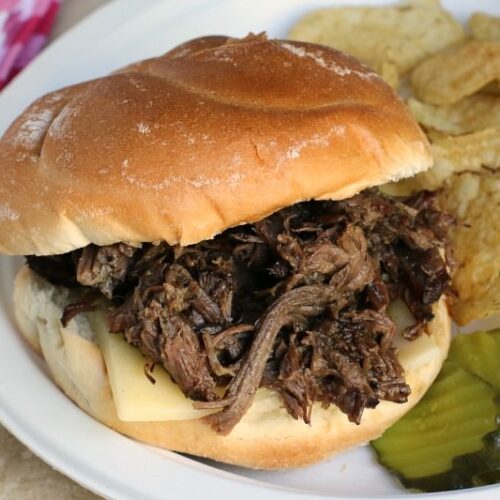 beef sandwich on plate with chips and pickles on side