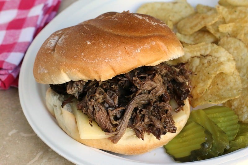 close up of beef sandwich on plate with chips and pickles on side