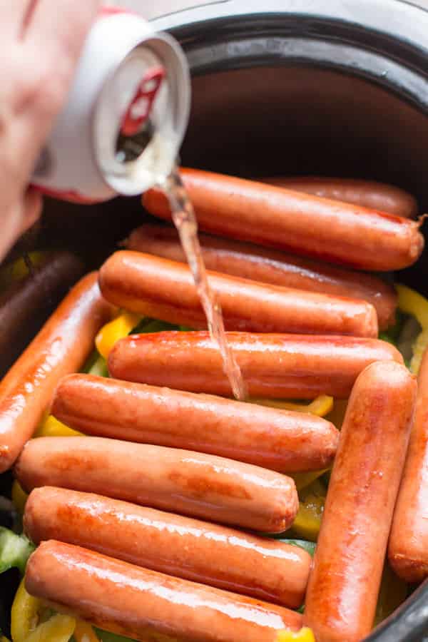 Beer being poured over browned brats in slow cooker.