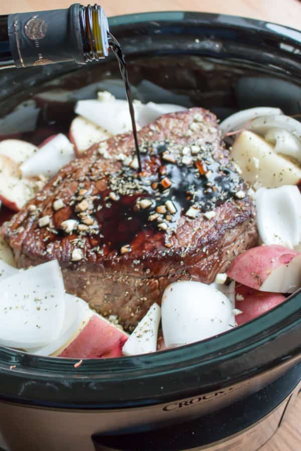 Balsamic vinegar being pour over beef roast, garlic and vegetables in slow cooker.
