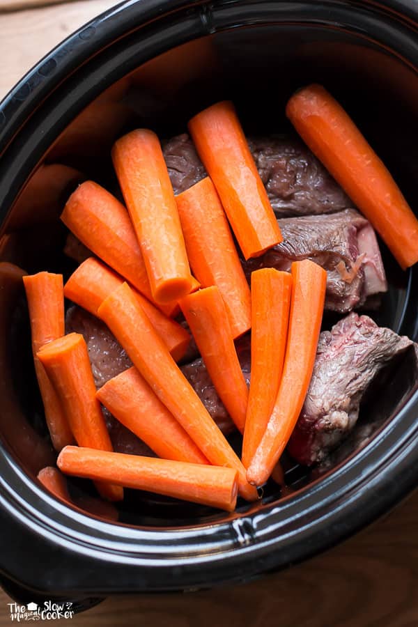 Slow cooker filled with short ribs with carrots on top.