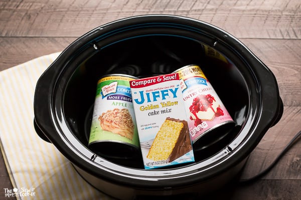 Cans of apple and cherry pie filling and box of jiffy yellow cake mix in slow cooker.