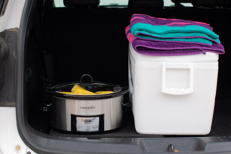 white cooler with towels on top and a slow cooker with corn on the cob in it.