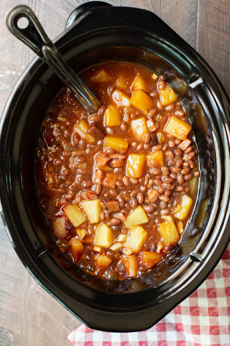 Baked beans in slow cooker with ham and pineapple chunks with red check napkin next to it.