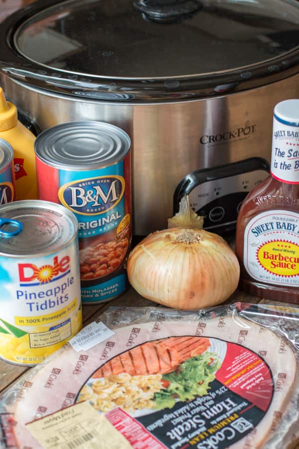 can of baked beans, mustard, barbecue sauce, ham steak and onion in front of slow cooker.