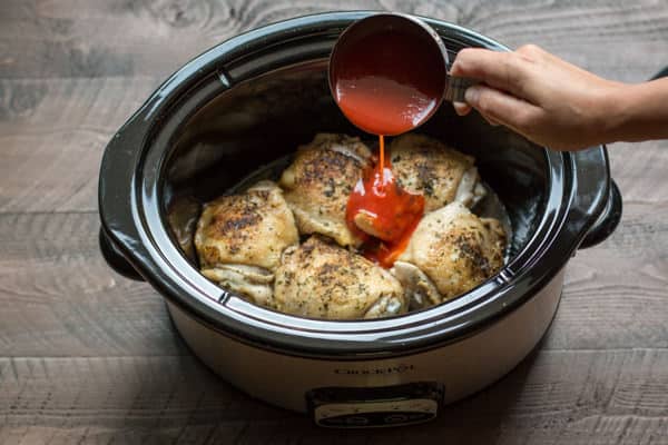 Chicken thighs in slow cooker with buffalo sauce being poured over.