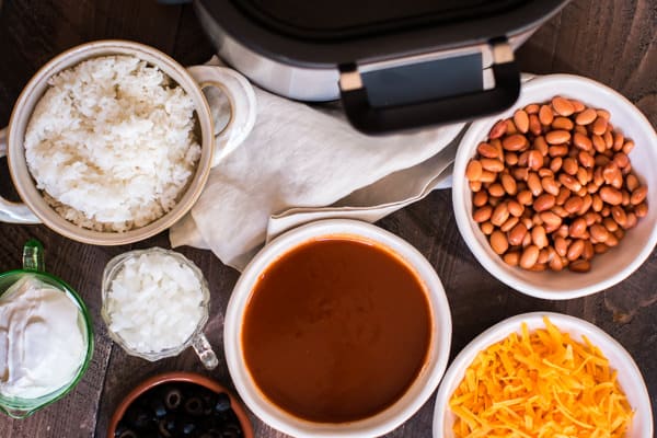 rice, onion. pinto beans, cheese, olives, ground beef and enchilada sauce next to a slow cooker.