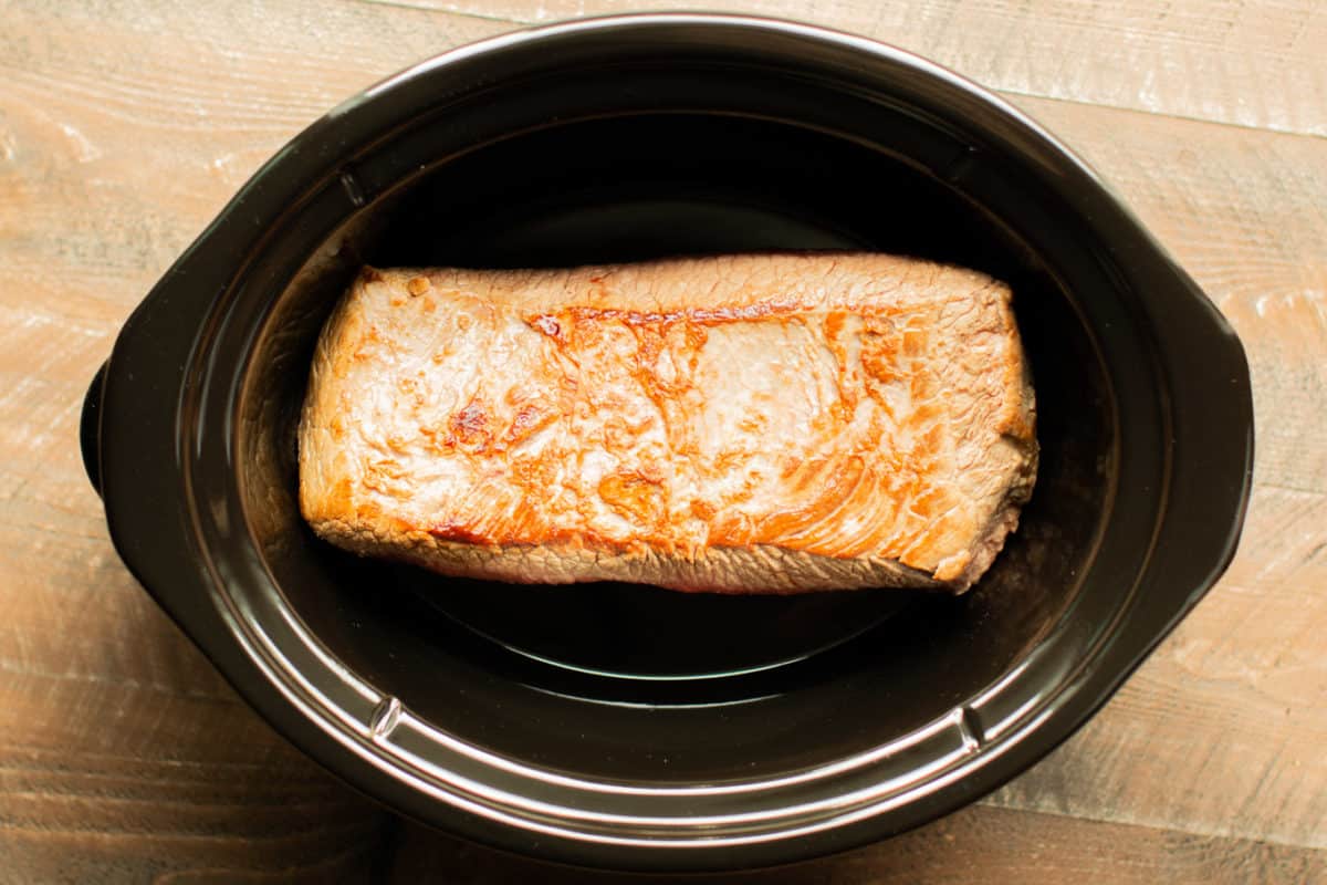 browned brisket alone in a black oval slow cooker.