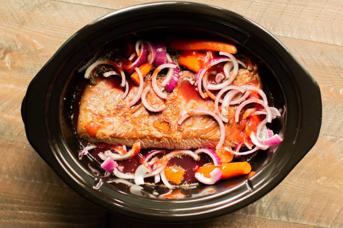 brisket in the slow cooker with raw red onion and carrots on top.