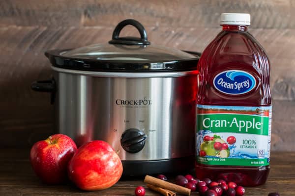cran apple juice, apples and cinnamon sticks in front of a slow cooker.