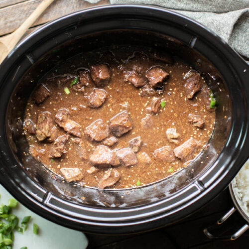 cooked teriyaki beef in the slow cooker with green onions and rice on the side.