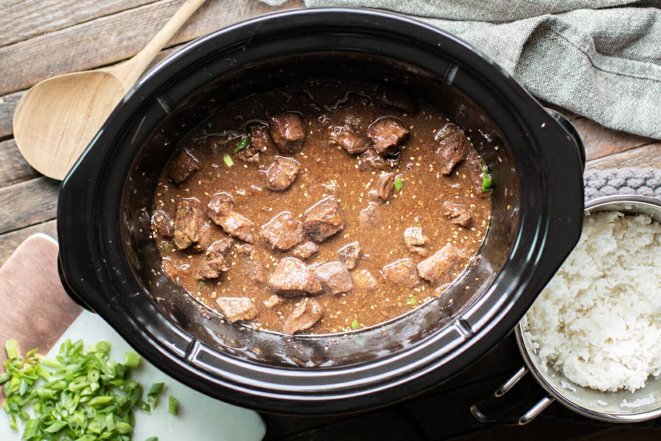 cooked teriyaki beef in the slow cooker with green onions and rice on the side.