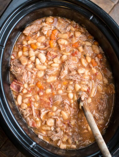 white beans and pork in a slow cooekr with a wooden spoon in it.