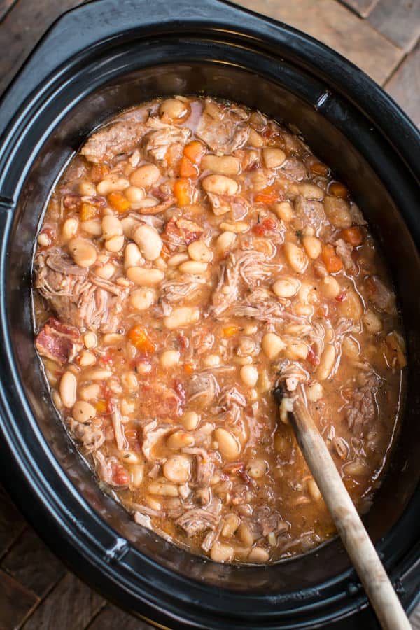 white beans and pork in a slow cooekr with a wooden spoon in it.
