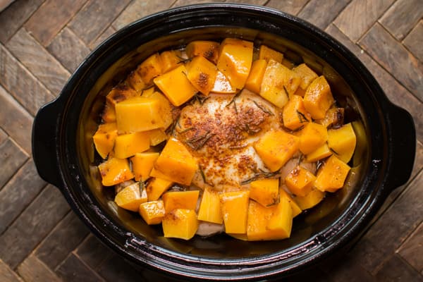 whole chicken in a slow cooker with butternut squash cubes, garnished with rosemary.