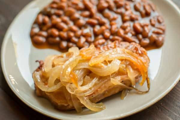 pork chop on a plate with onions on top. Baked beans on the side.