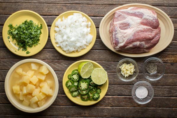 pork roast, onion, pineapple, jalapeno, line, garlic and cilantro on a wood table.