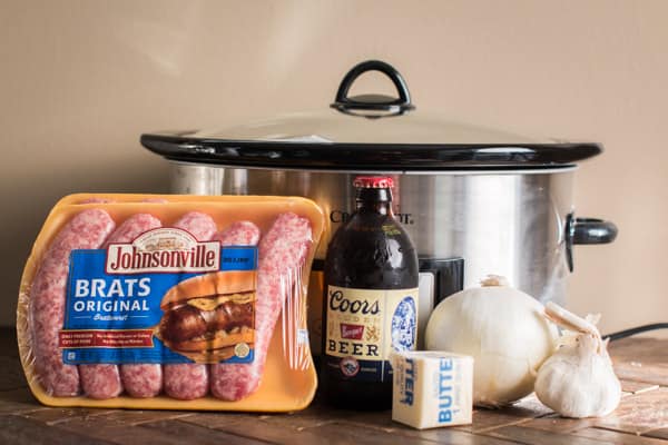 raw brats, coors beer, butter, onion and garlic in front of a slow cooker