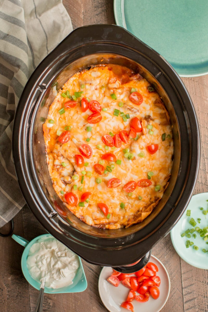 Enchilada casserole in slow cooker with tomatoes and green onion on top.