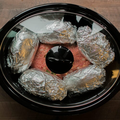 raw meatloaf in slow cooker with potatoes wrapped in foil on top.