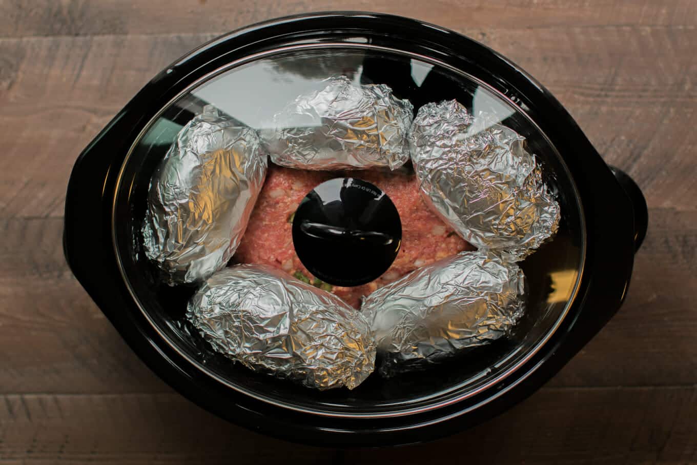 raw meatloaf in slow cooker with potatoes wrapped in foil on top.