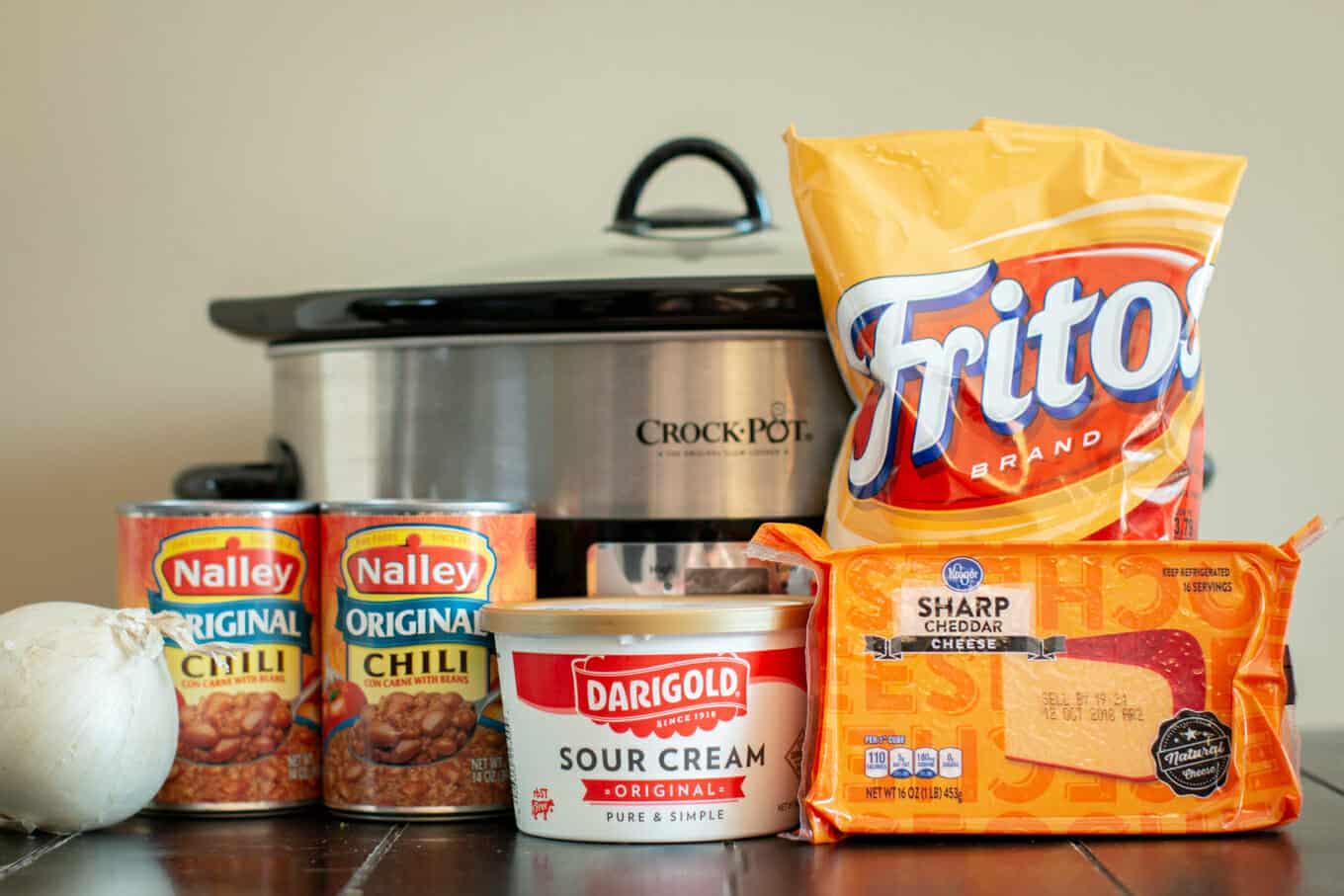 cans of chili, sour cream, fritos, cheese, and onion in front of a slow cooker.