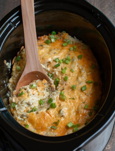 cheesy potatoes in a slow cooker with a wooden spoon in it.