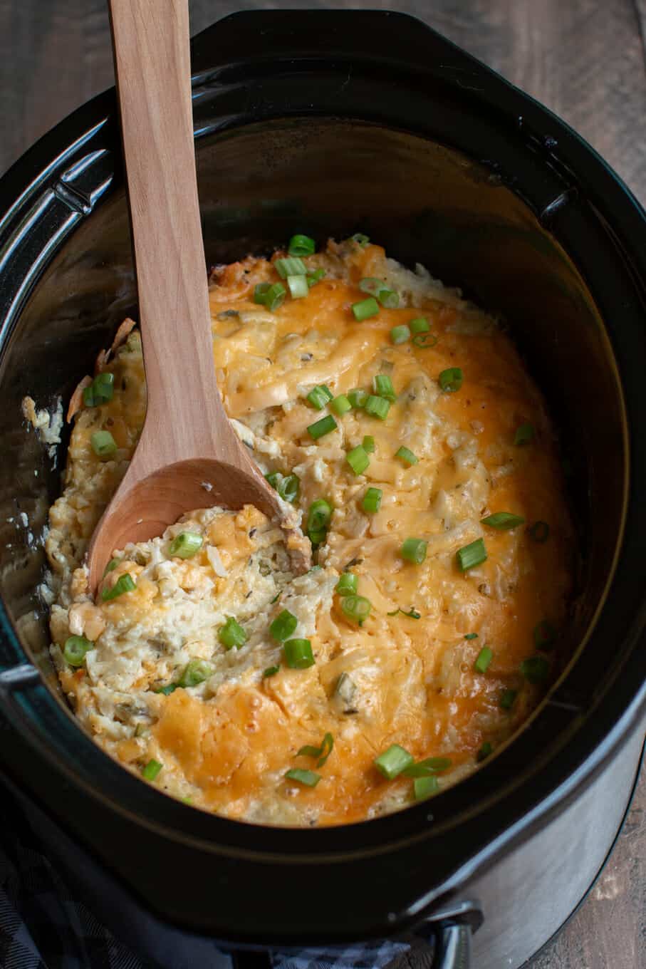 cheesy potatoes in a slow cooker with a wooden spoon in it.