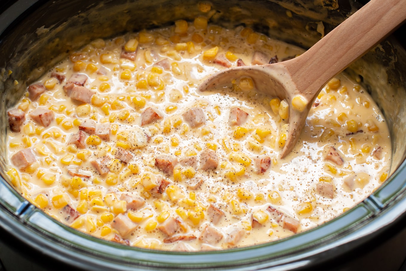 Wooden spoon serving ham and cheese corn from a slow cooker.